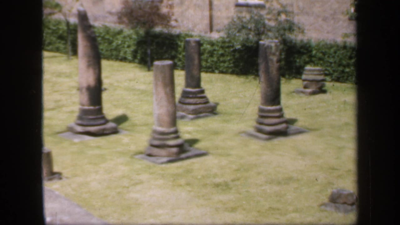 Stone columns and low ruins on a grassy courtyard seen through dark window frames.