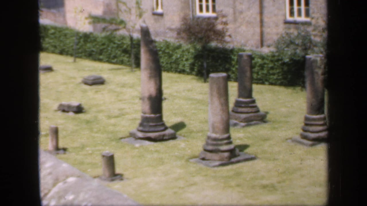 1949 England 8mm view of upright stone columns and small ruin fragments on grass beside a hedge and masonry building.