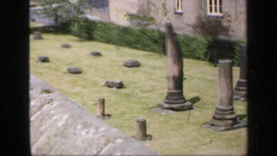 1949 England 8mm frame looking down over a roof edge toward stone columns and scattered ruin fragments in a lawned courtyard.