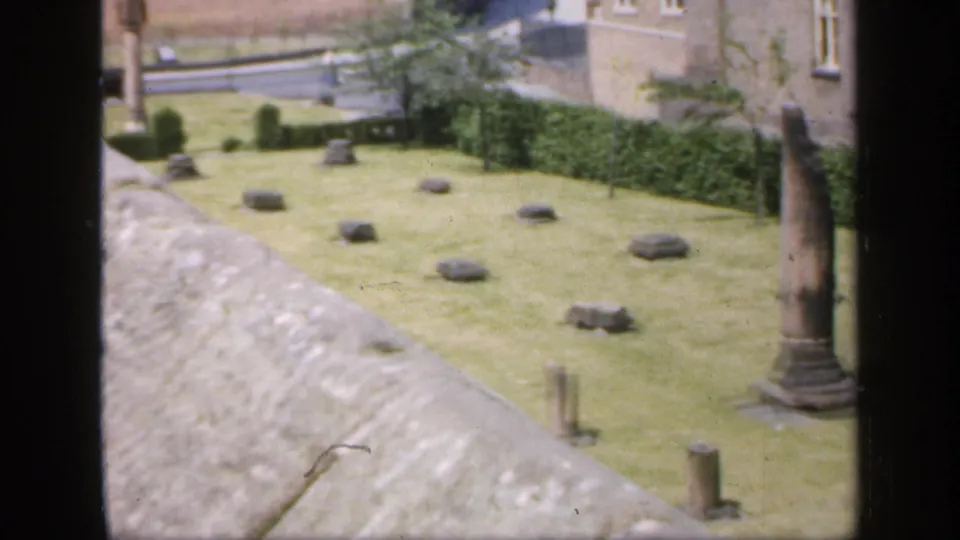 1949 England 8mm frame showing a wider garden-ruins view with stone columns, low stones, hedges, and a road beyond the wall.