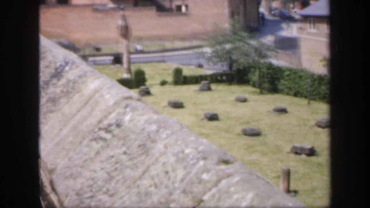 1949 England 8mm frame showing a stone parapet above a grassy historic courtyard with scattered markers, a monument, and brick buildings.