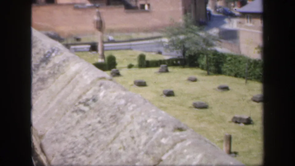 1949 England 8mm frame showing a stone parapet above a grassy historic courtyard with scattered markers, a monument, and brick buildings.