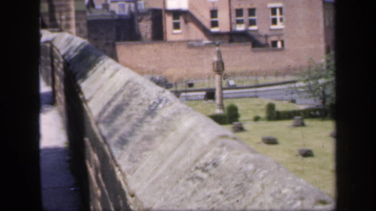 Stone parapet and walkway beside a grassy courtyard with buildings in the background.