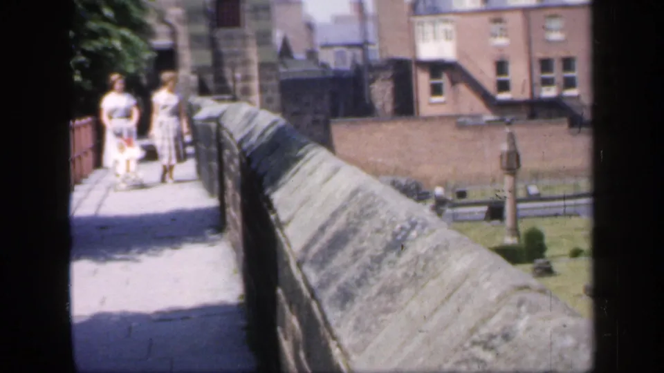 Two women in light dresses walk along a stone parapet walkway in 1949 England, with courtyard buildings and grass visible behind them.