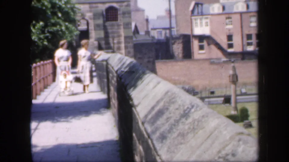 A 1949 England home-movie frame shows two women walking beside a stone parapet, with a grassy courtyard and brick buildings in the distance.