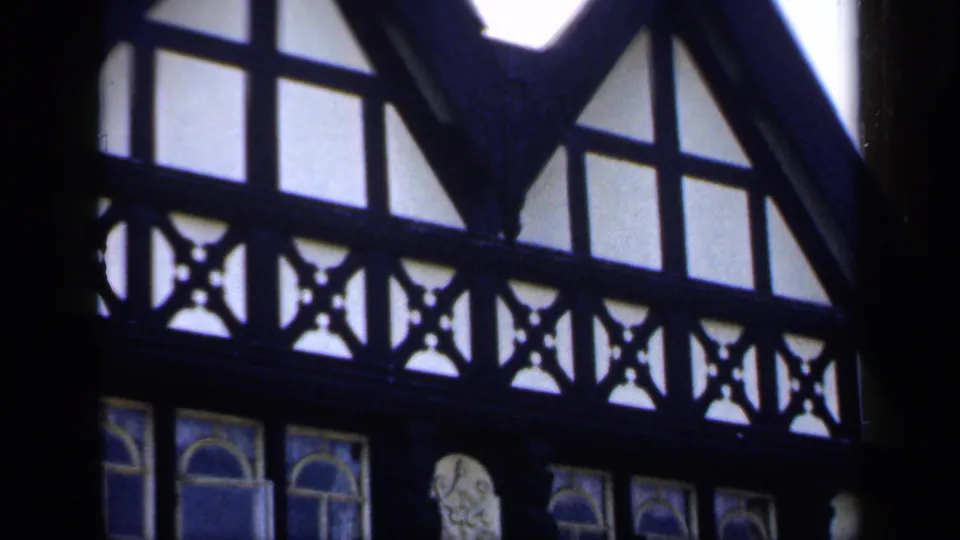 1949 England 8mm frame showing dark timber framing, high interior windows, and decorative arched glass inside a church-like space.