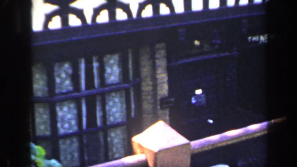 1949 England 8mm frame showing a dark doorway, brick wall, lower windows and a railing or ledge in the foreground.