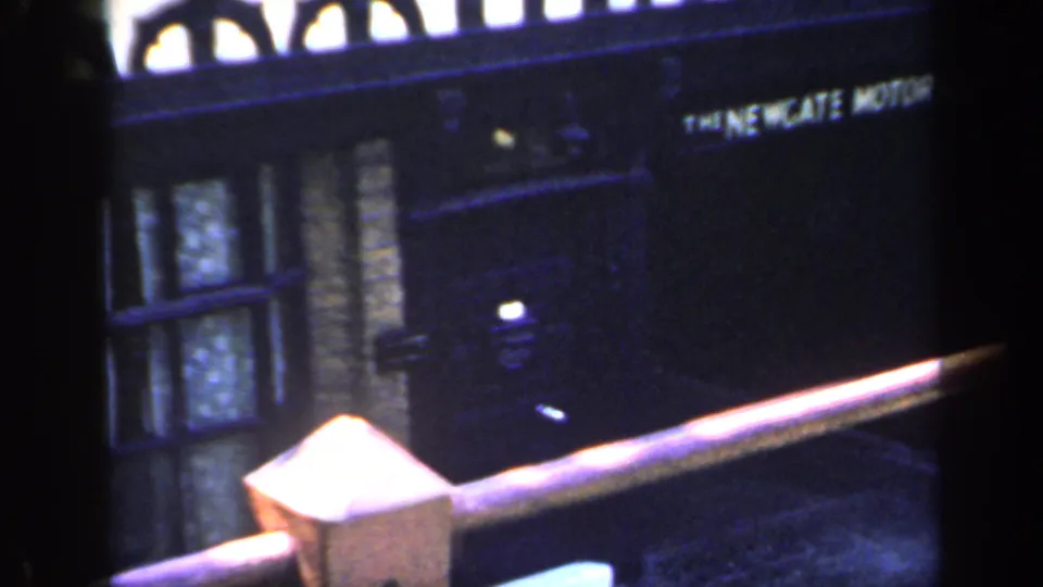1949 England 8mm frame showing a shaded view toward brick frontage with a motor company sign and dark foreground obstruction.