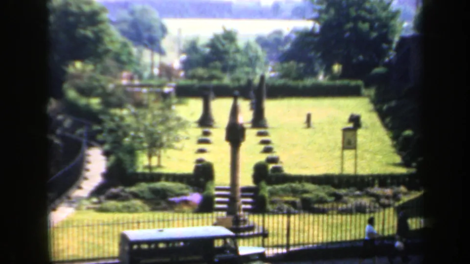 1949 England 8mm view of a manicured public garden with a stone monument, iron fence, shrubs, distant trees, and a bus at the curb.