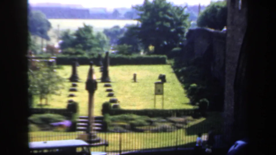1949 England home-movie frame showing a formal park lawn, central stone monument, clipped hedges, trees, railings, and street activity nearby.
