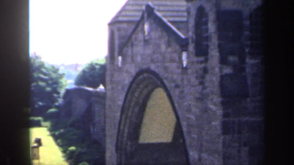 1949 England frame with Gothic-style stonework, an arched opening, hedges, railings, and part of the garden visible at left.