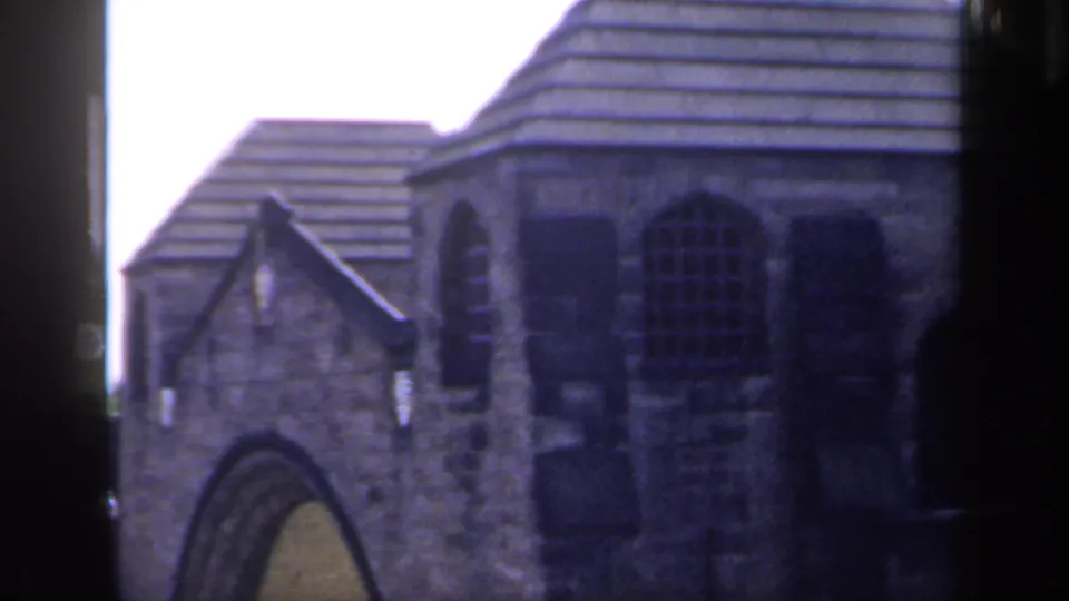 1949 England 8mm frame showing the upper stone building, tiled rooflines, arched window, and shaded doorway area.