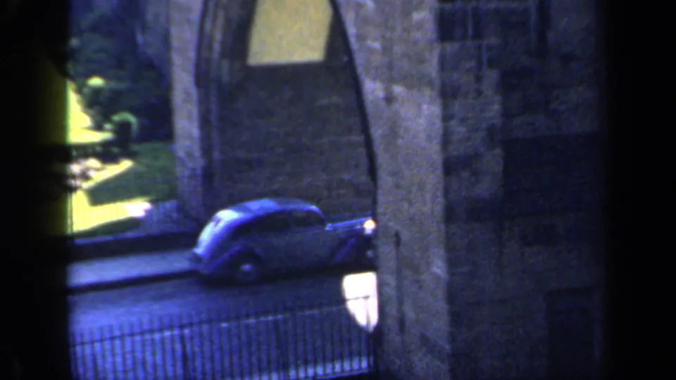 1949 England 8mm frame showing a blue period car moving along the street beside a stone arch, railing, and shaded building edge.