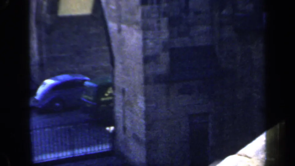 1949 England 8mm frame with a blue period car beneath a stone arch, framed by dark foreground edges and gridded windows.