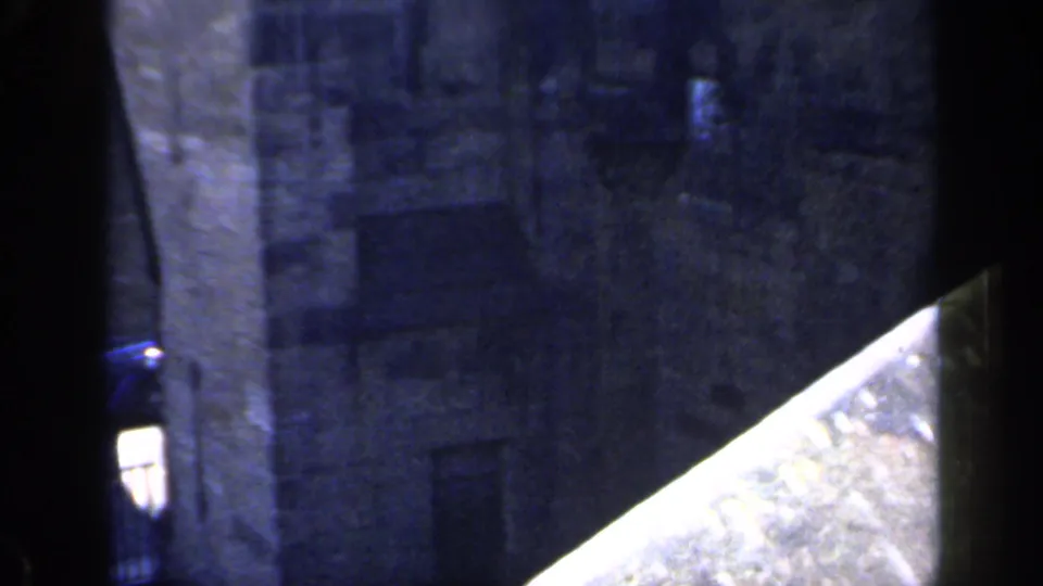 1949 England home-movie frame with a grey castle wall, dark vertical frame edges, and a bright angled stone surface in the foreground.