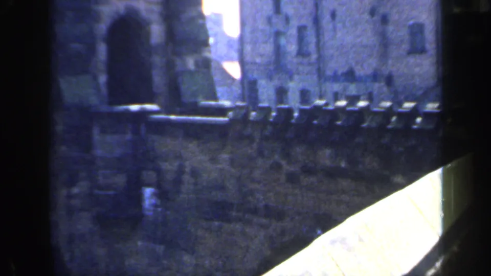 1949 England home-movie frame showing an arched castle entrance, grey stone wall, parapet, and narrow walkway.