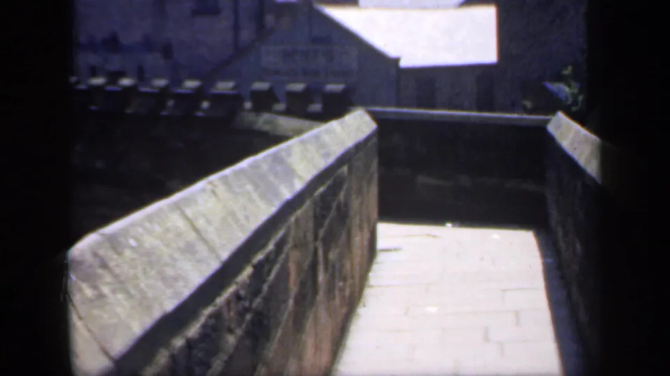 1949 England home-movie frame showing the walkway closer to the camera, with sunlit paving and a stone wall beside it.