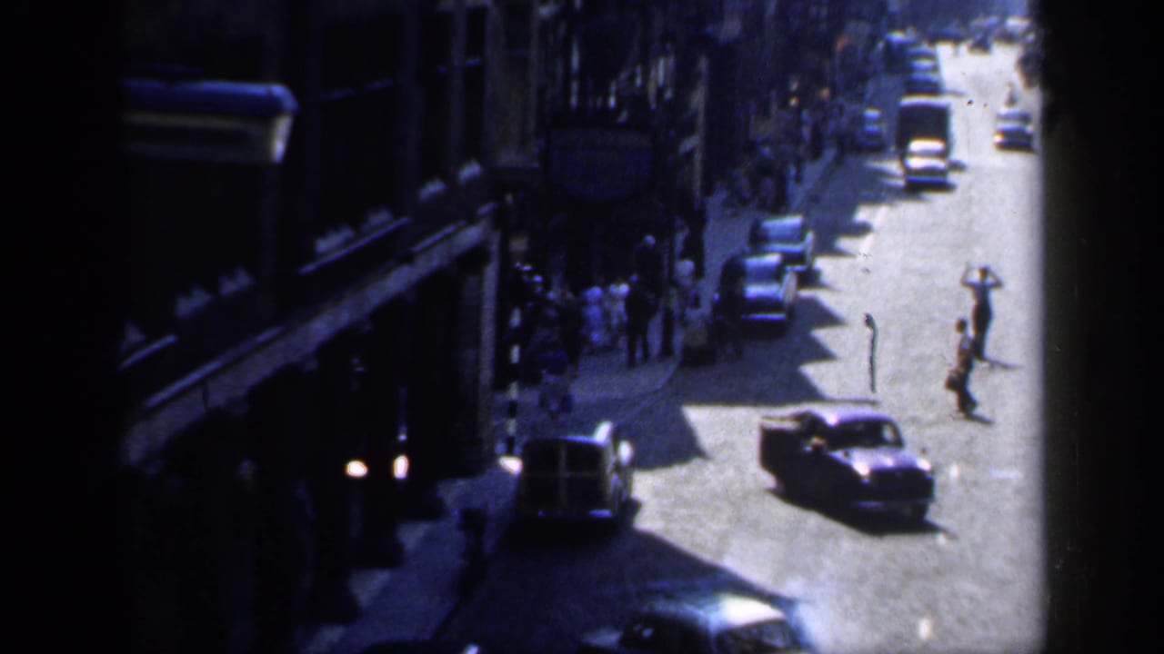 High-angle 1949 England street view with pedestrians crossing, period cars, and shopfronts in strong sun and shadow.