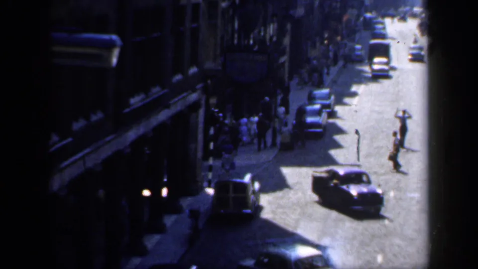 High-angle 1949 England street view with pedestrians crossing, period cars, and shopfronts in strong sun and shadow.