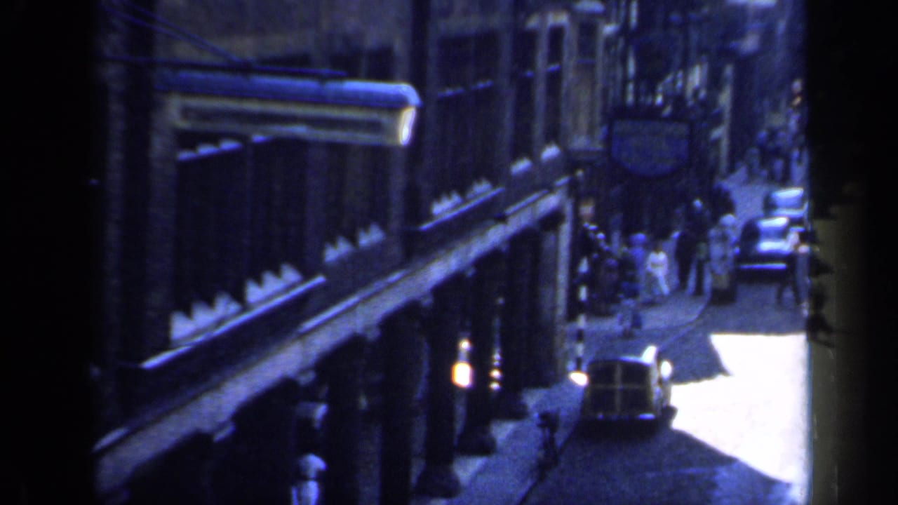 Elevated 1949 England view of a street edge with pedestrians near shops, a parked car, and strong sunlight beyond the shadowed frontage.
