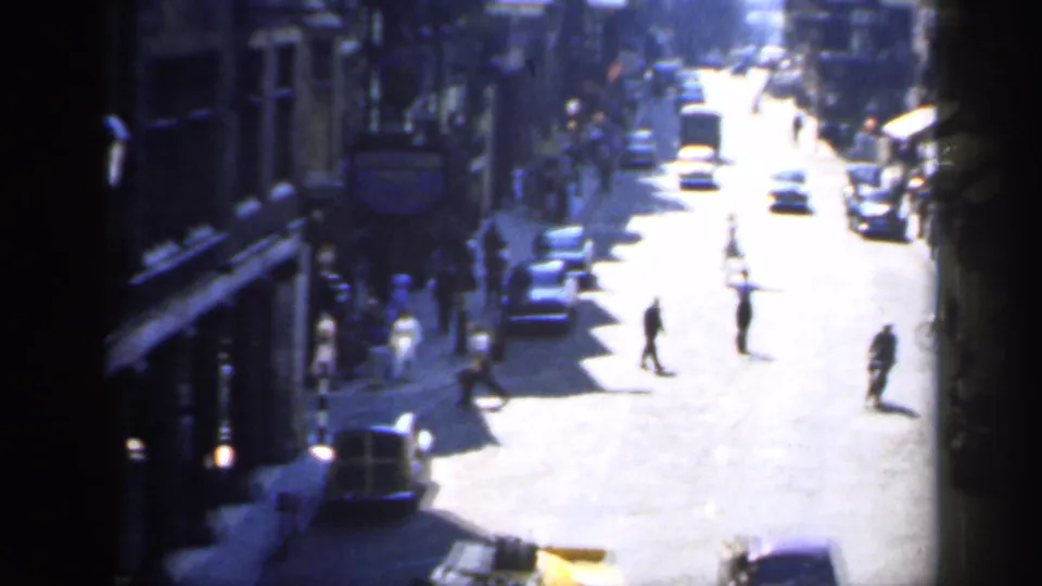 Sunlit 1949 England city street with several pedestrians crossing between period cars and shaded shopfronts.