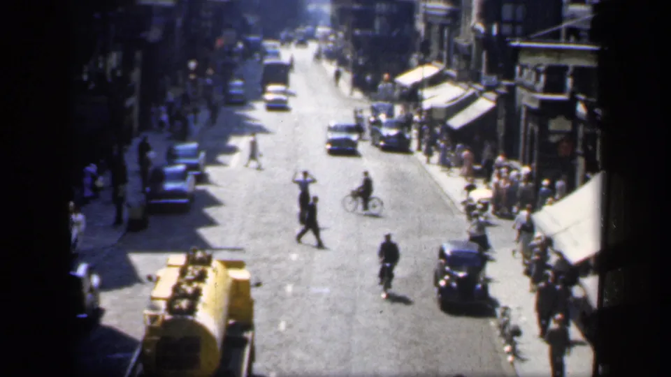 High-angle 1949 England street scene with a cyclist, pedestrians, period cars, storefront awnings, and a yellow tanker truck.