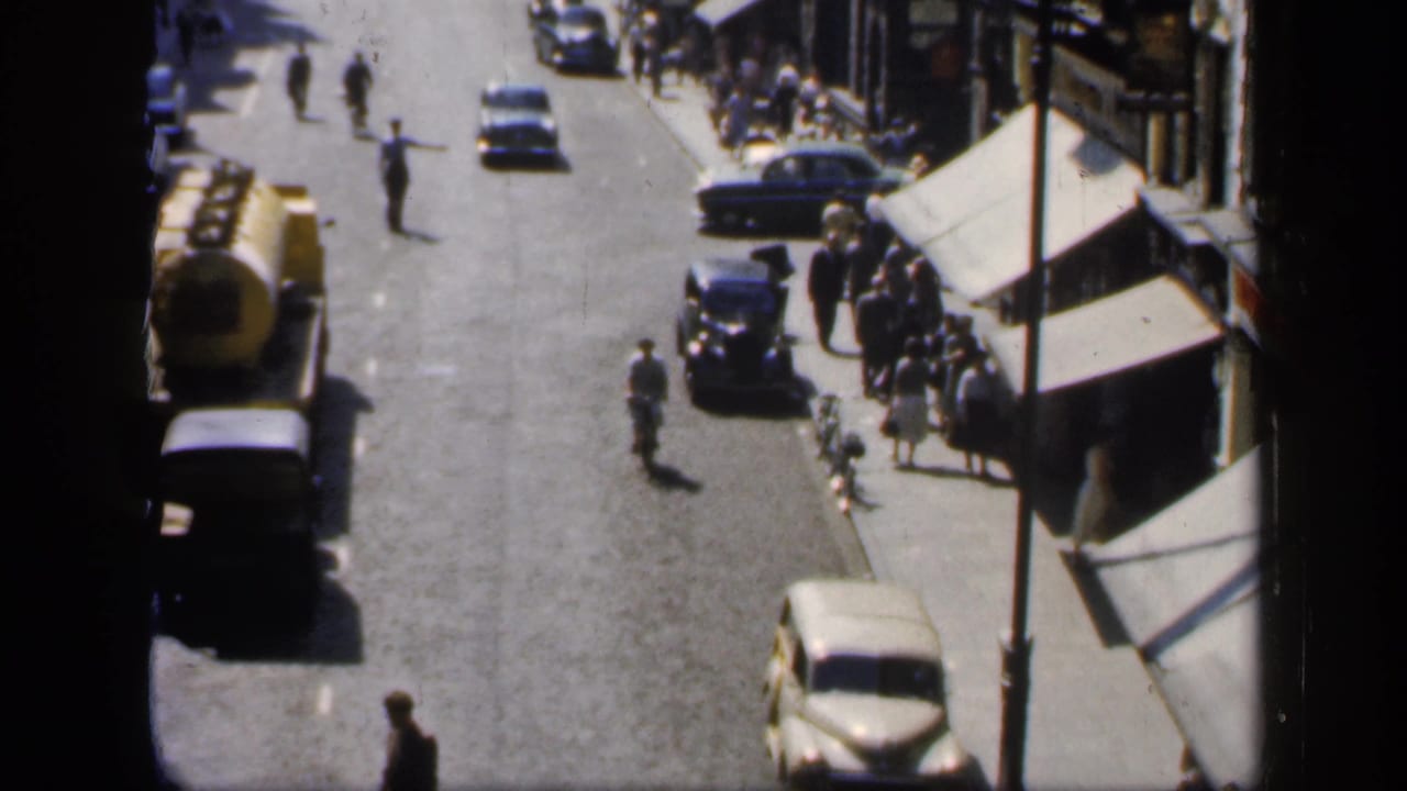 High-angle street view with cars, a yellow tanker truck, pedestrians, and shop awnings.