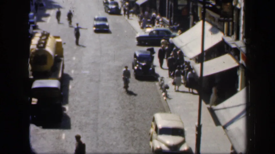 High-angle 1949 England street scene with classic cars, a yellow tanker-style lorry, pedestrians, and shopfront awnings.
