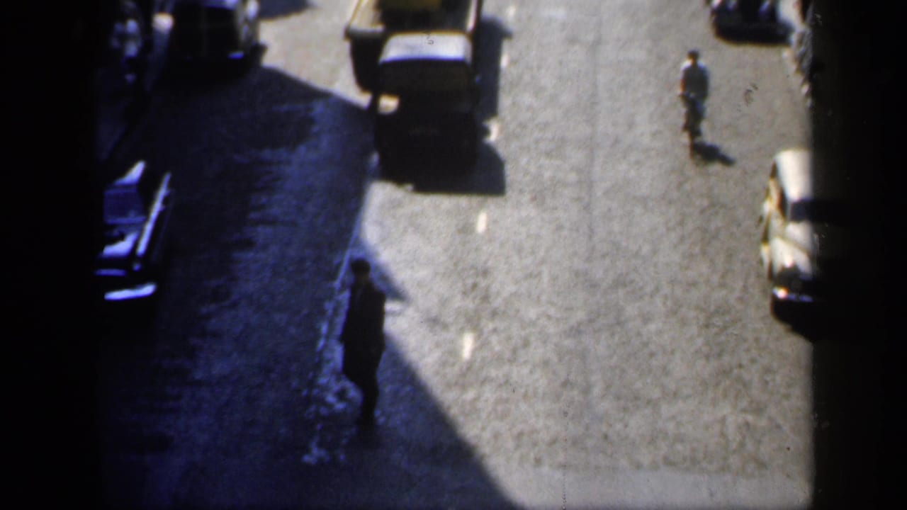 High-angle view of a 1949 England street with a dark car near deep shadow and pedestrians moving along the road.