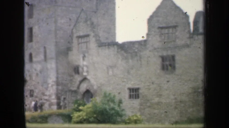 1949 England home-movie frame showing a stone ruin facade with arched doorway and small distant visitors near the wall.