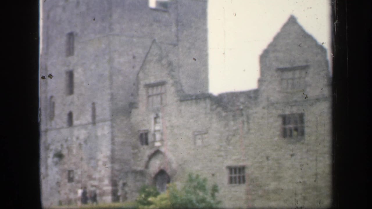 Stone castle facade with tower, arched doorway, windows, and visitors near the base.