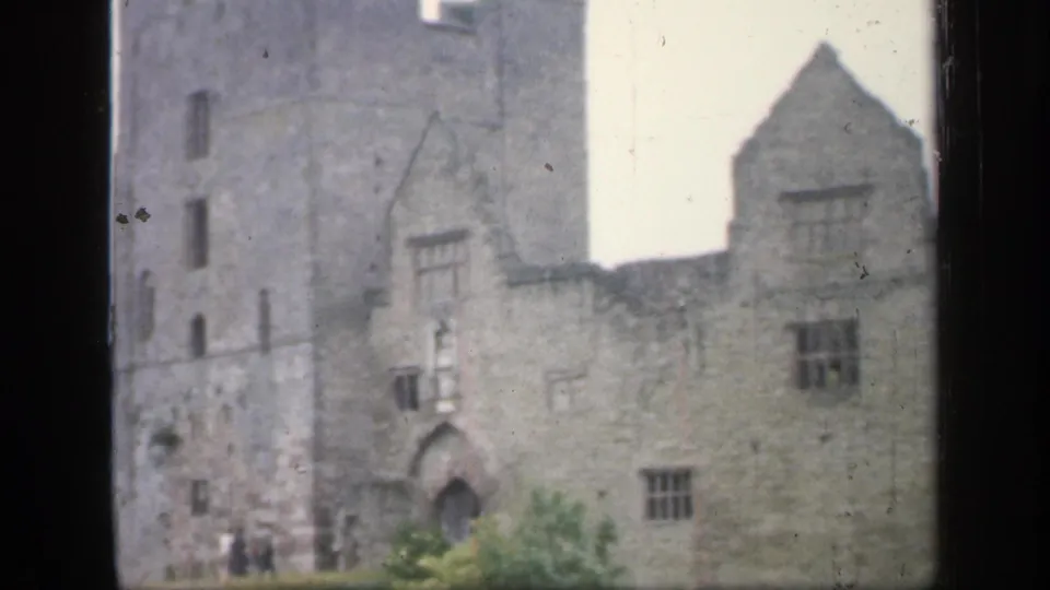 1949 England 8mm frame showing a large stone castle or fortified building with a tower, arched entrance, and distant visitors.