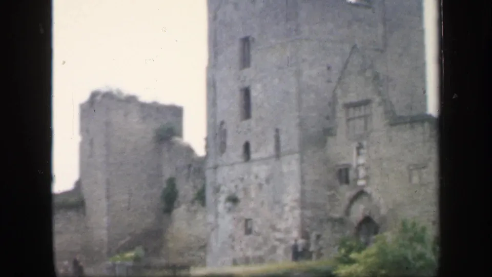 1949 England home-movie frame showing a stone tower beside a lower ruined wall, with visitors gathered near the base.
