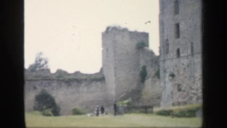 1949 England 8mm frame showing castle grounds with a ruined wall, square stone tower, lawn, shrubs, and distant visitors.