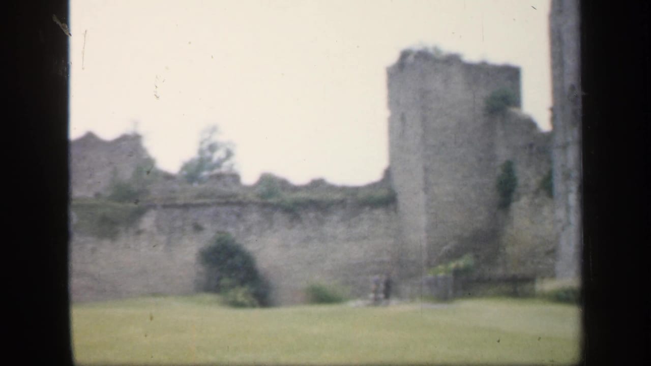 1949 England 8mm frame showing weathered castle walls and square towers across an open lawn, softly focused and partly framed by dark gate edges.