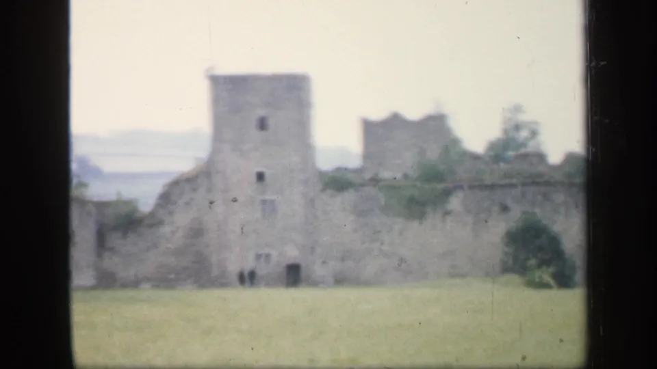 1949 England home-movie frame of a square tower and crumbling wall, with small indistinct visitors at the base and muted color from 8mm film.