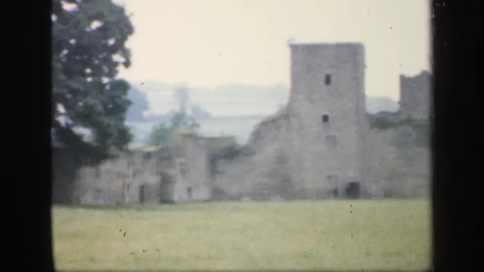 Soft 8mm frame of English castle ruins, showing a square tower, low wall sections, a large tree, open lawn, and pale distant landscape.