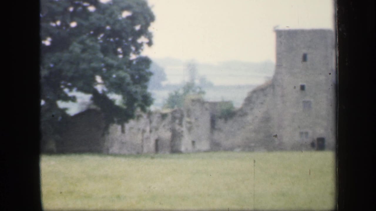 Stone castle ruins and a square tower beyond a green field, framed by dark film borders.