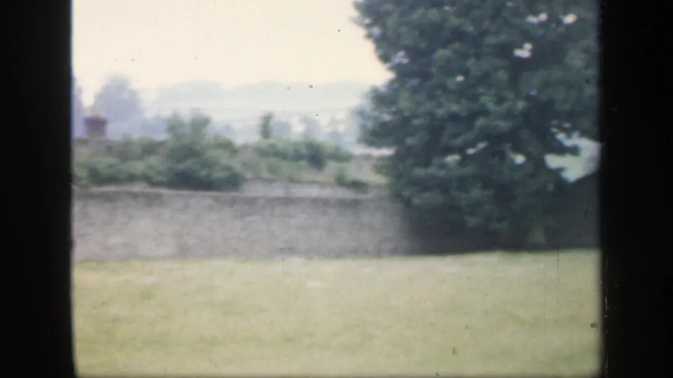 1949 England 8mm frame looking past pasture and a low ruin wall toward trees and hazy countryside.
