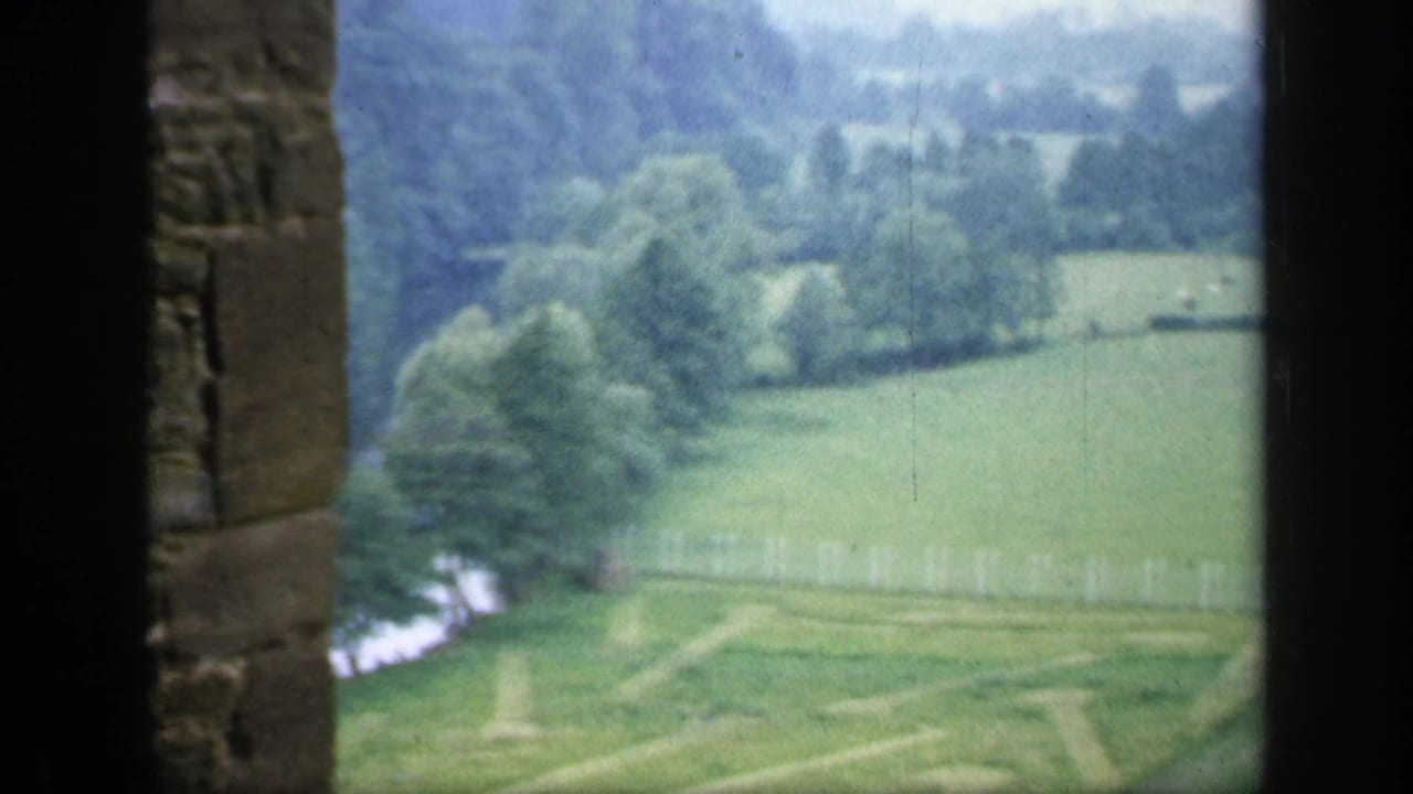 Green countryside seen through a dark stone window opening with fields, trees, and a pale fence.