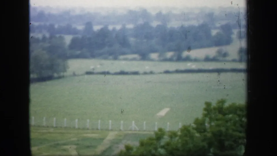 1949 England 8mm frame of a green countryside view, with distant trees, pale fencing, and dark window edges surrounding the scene.