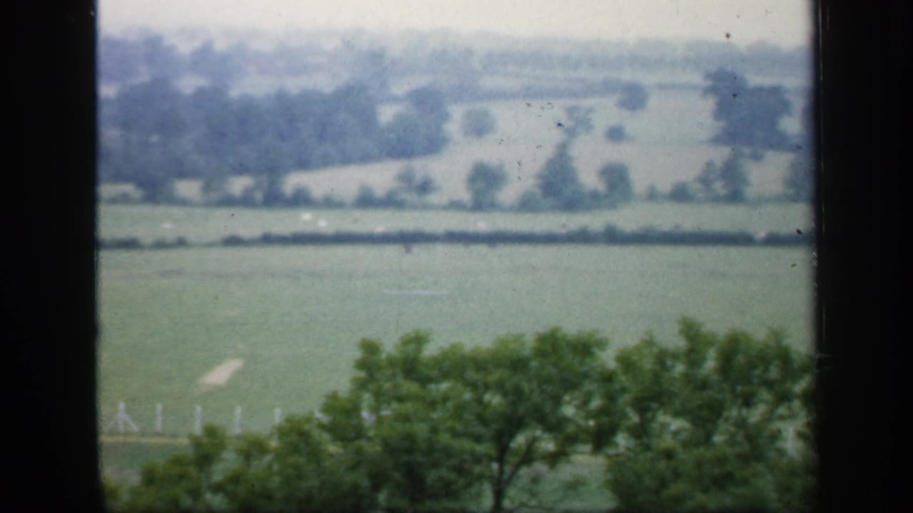 Soft 8mm view of English farmland in 1949, with hedgerows, trees, and a dark vertical window frame in the foreground.