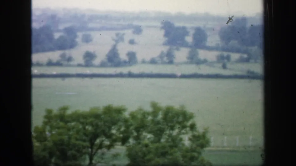 Muted 1949 home-movie frame of English fields beyond foreground trees, with hedgerow lines and film dust visible.
