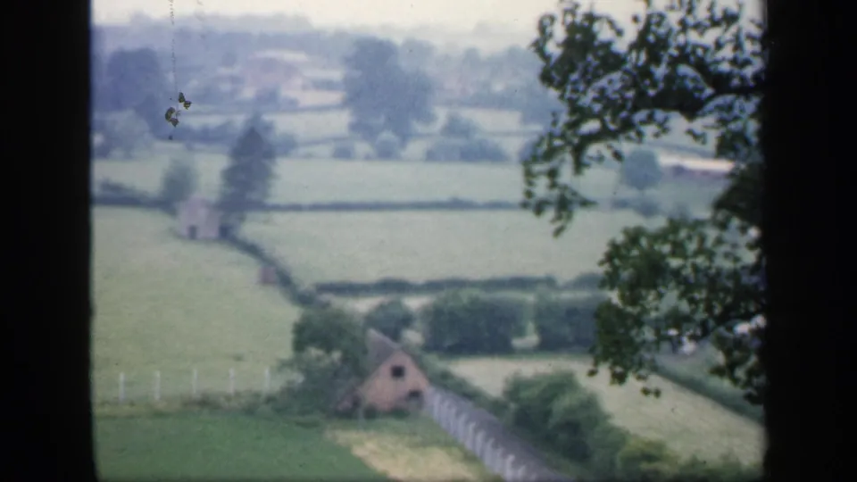 1949 England home-movie frame showing a small rural building below hedged fields, pale fence posts, and leafy branches.