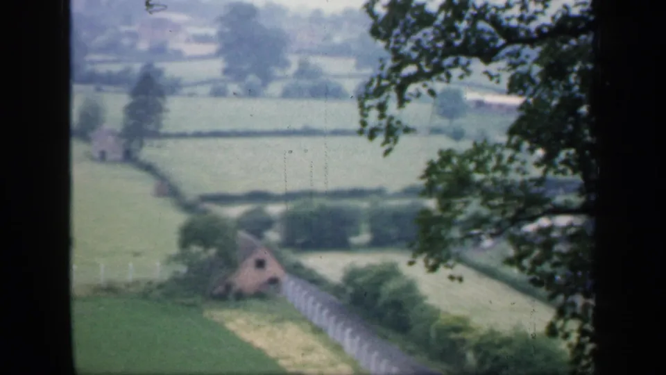1949 England countryside frame with a small farmhouse or outbuilding, pale fence line, hedgerows, fields, and overhanging branches.
