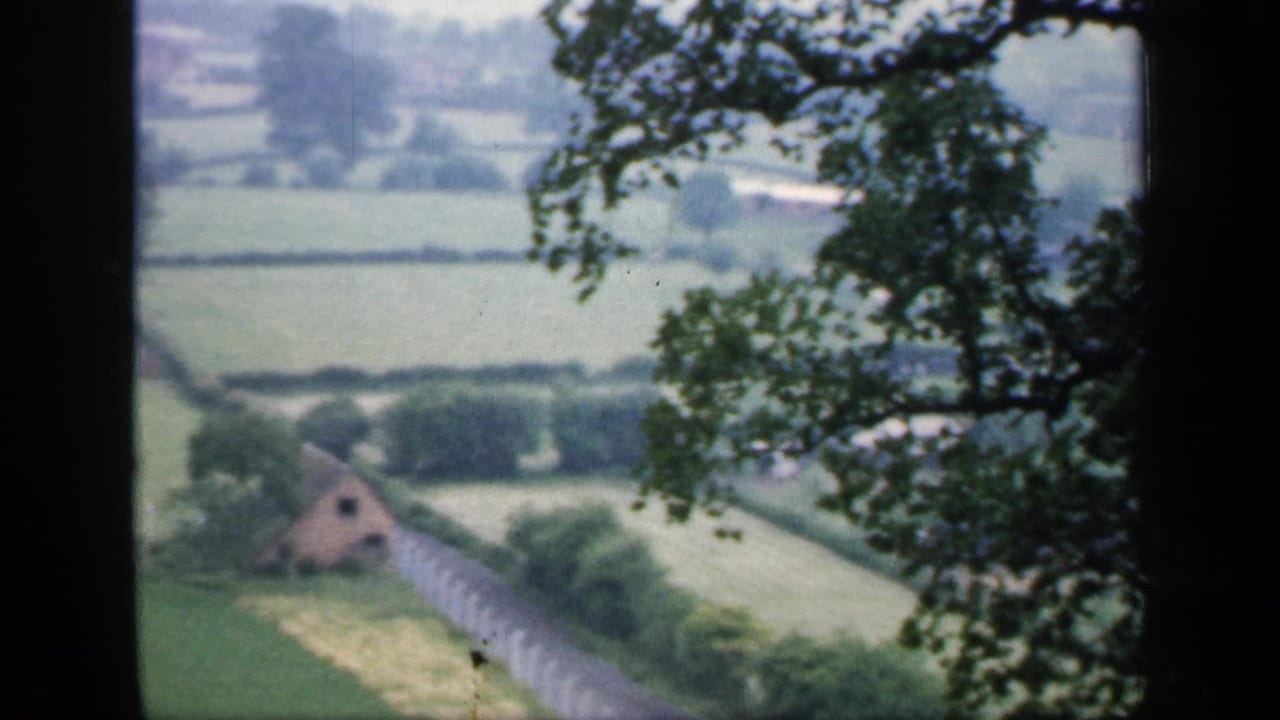 1949 England home-movie view of green fields, hedgerows, a pale fence, and a small distant building framed by trees.
