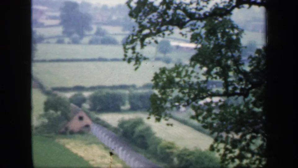 1949 England home-movie view of green fields, hedgerows, a pale fence, and a small distant building framed by trees.