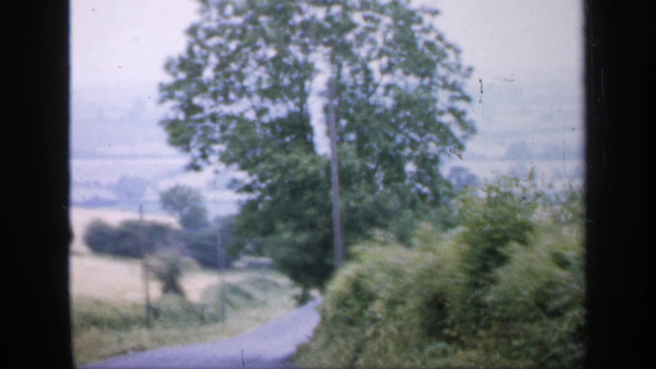 1949 England home-movie frame from a moving vehicle, showing a narrow country road, hedgerows, trees, and distant fields.