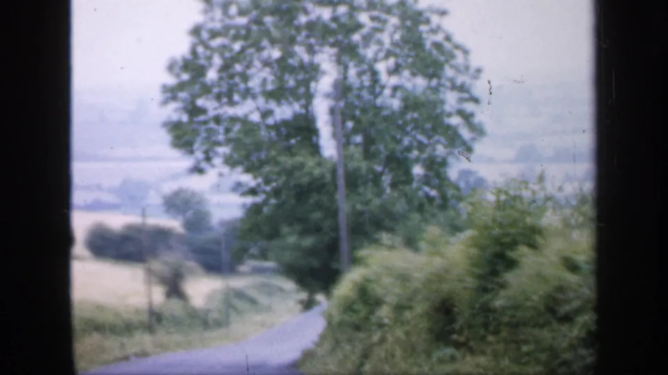 1949 England home-movie frame from a moving vehicle, showing a narrow country road, hedgerows, trees, and distant fields.