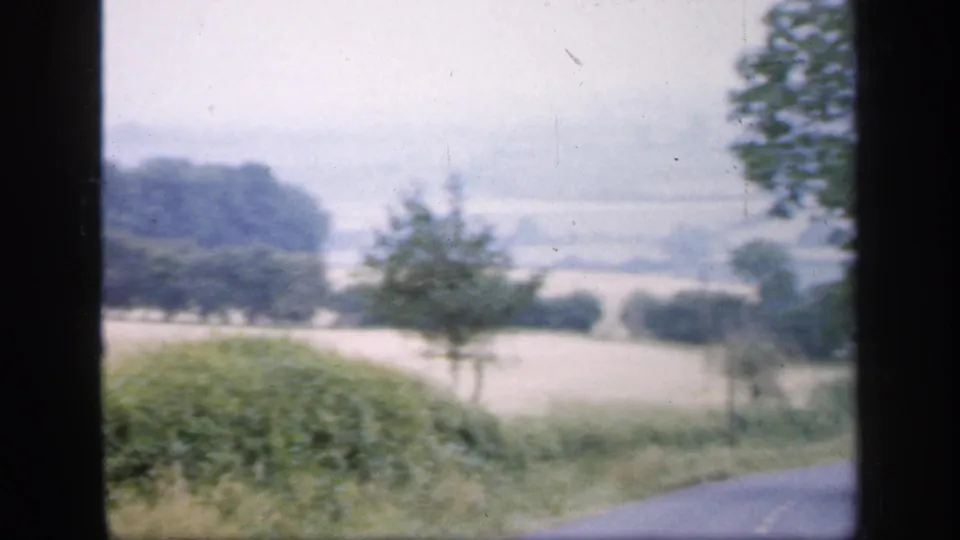 1949 English countryside seen through 8mm film grain, with hedgerows, pale fields, and distant trees from a moving viewpoint.